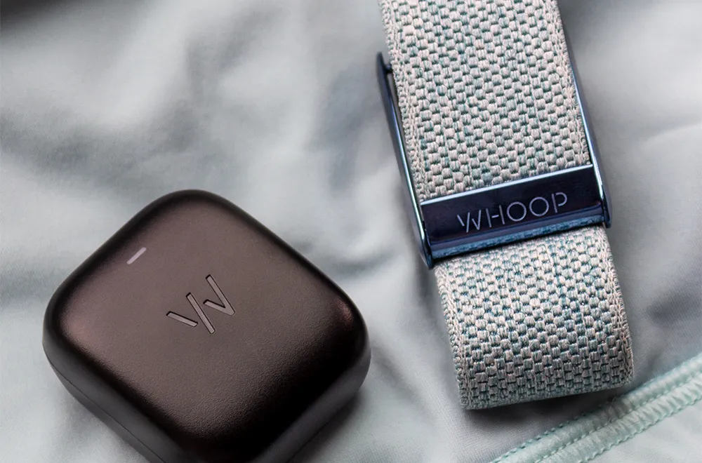 A WHOOP fitness band on a wrist next to a smartphone showing recovery and strain metrics, with a soft Gulf skyline backdrop