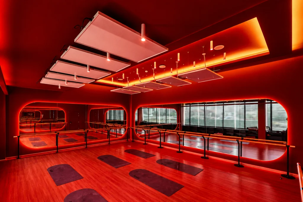 Red-lit group fitness studio at The Revive Club with rows of exercise mats on a wood floor, mirrored walls with rounded neon-edged panels, ballet barres, and acoustic ceiling panels.
