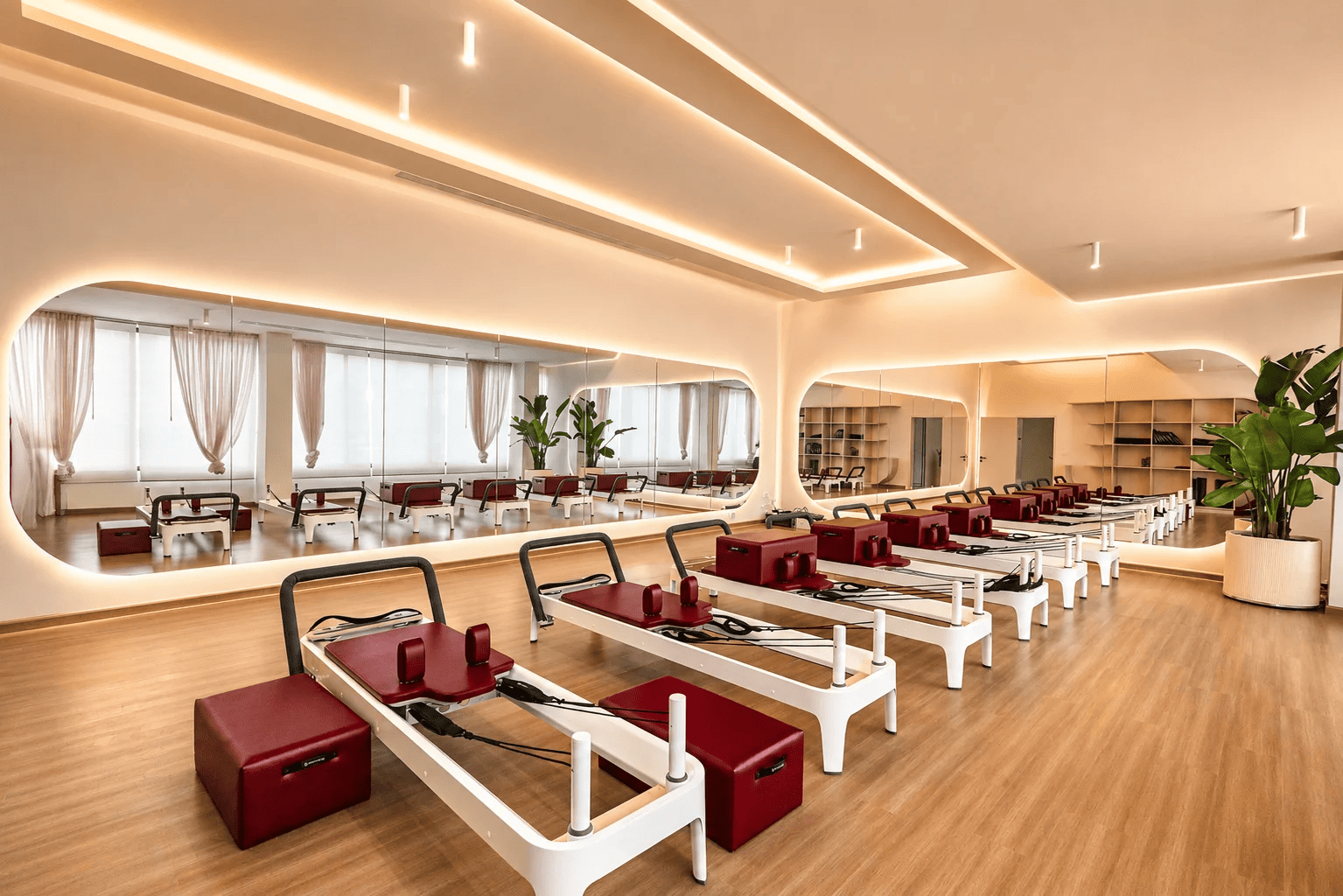 Bright, neutral-toned Pilates studio at The Revive Club with a row of reformer machines featuring burgundy upholstery, mirrored walls with backlit rounded panels, draped windows, and potted plants.