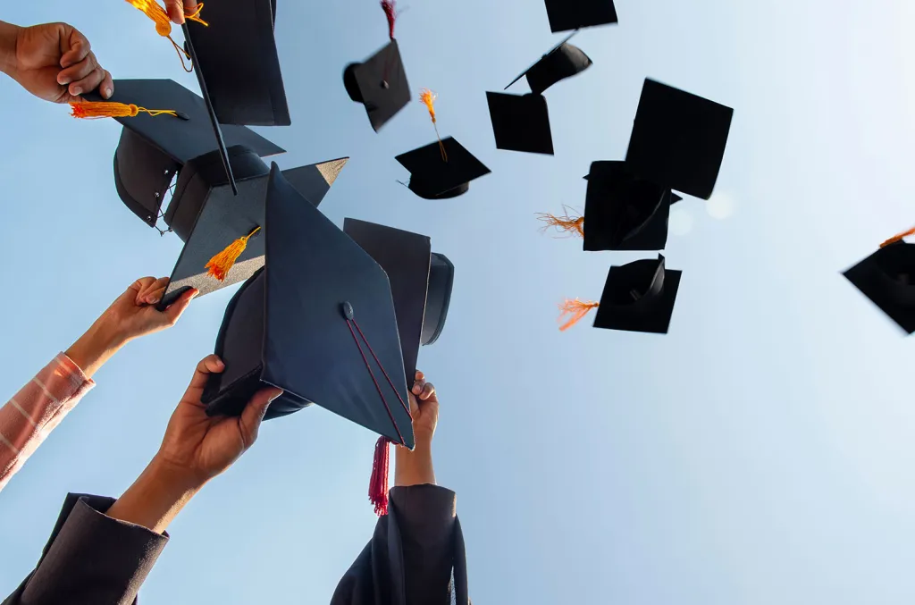 A Lebanese university graduate in a doctoral cap and gown stands in front of a Beirut campus building, holding a rolled diploma.