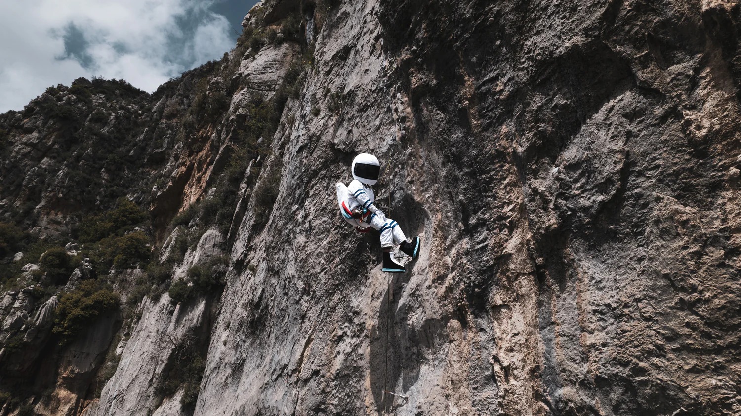 A climber wearing a white astronaut suit and helmet rappels down a rocky cliff face. A small Lebanese flag sits in the top-left corner. 