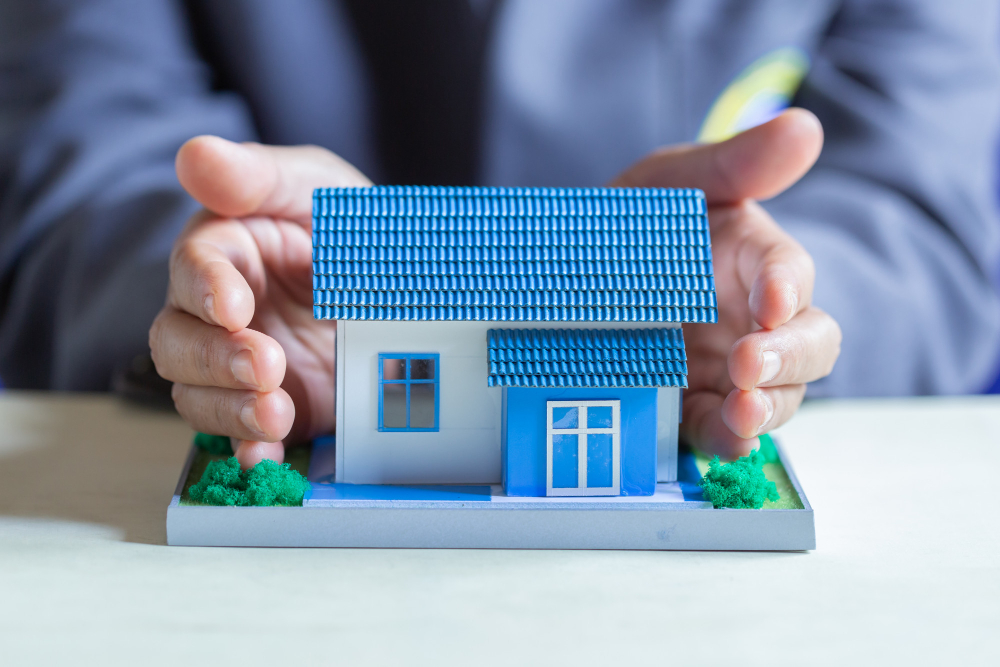 Hands cupped protectively around a small model house with a blue tiled roof on a tabletop, symbolizing home ownership and housing loan protection in Lebanon