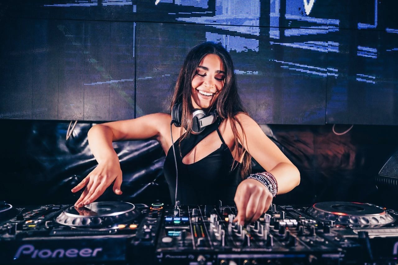 Lebanese DJ Chloe Kteily mixing on CDJ players behind a lit DJ booth, hands on the decks during a live set