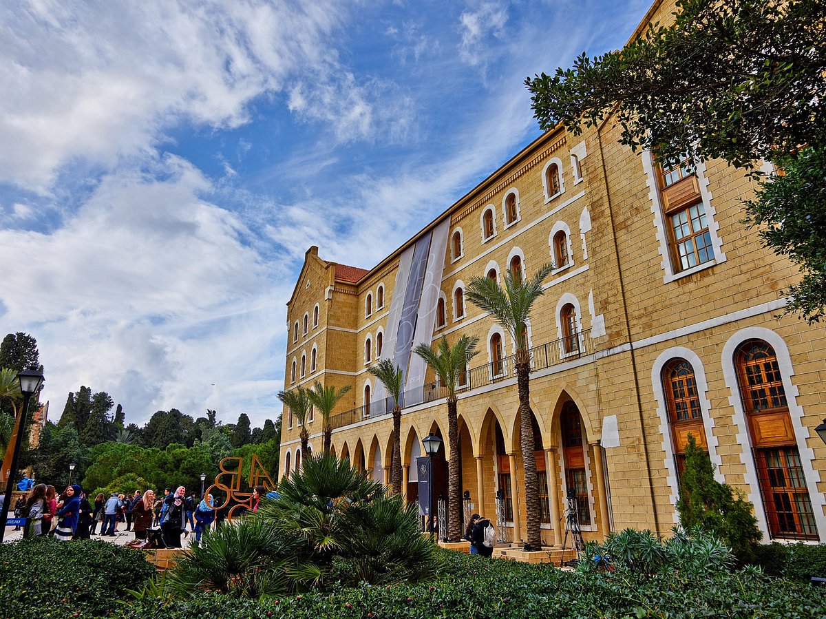 A sunny view of the historic yellow sandstone College Hall building on the American University of Beirut campus, with arched walkways, tall palm trees, green gardens, and students gathered near the entrance under a blue sky.
