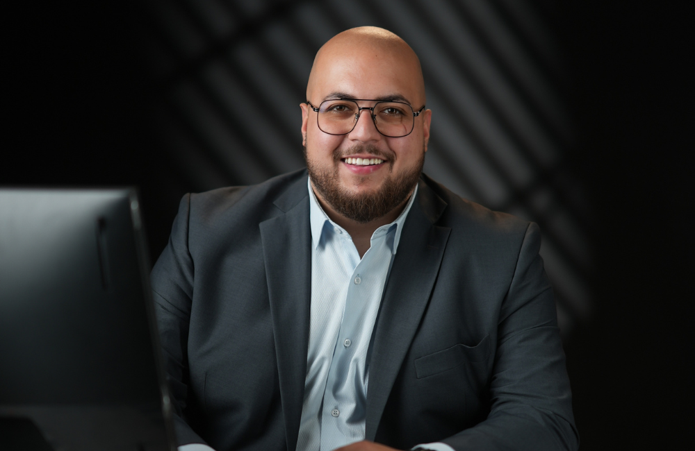Ali Haddad, Media Specialist at ZXCM, smiling in a dark suit and light shirt seated at his desk against a dark studio background.