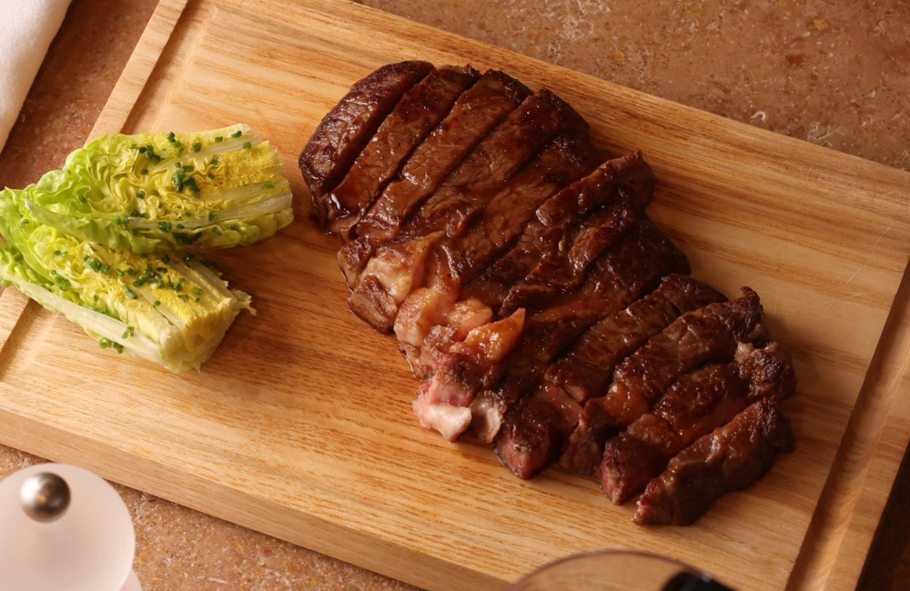 A sliced grilled steak on a wooden cutting board, served beside a halved baby gem lettuce topped with chives, with a salt grinder and glass of red wine in the foreground.