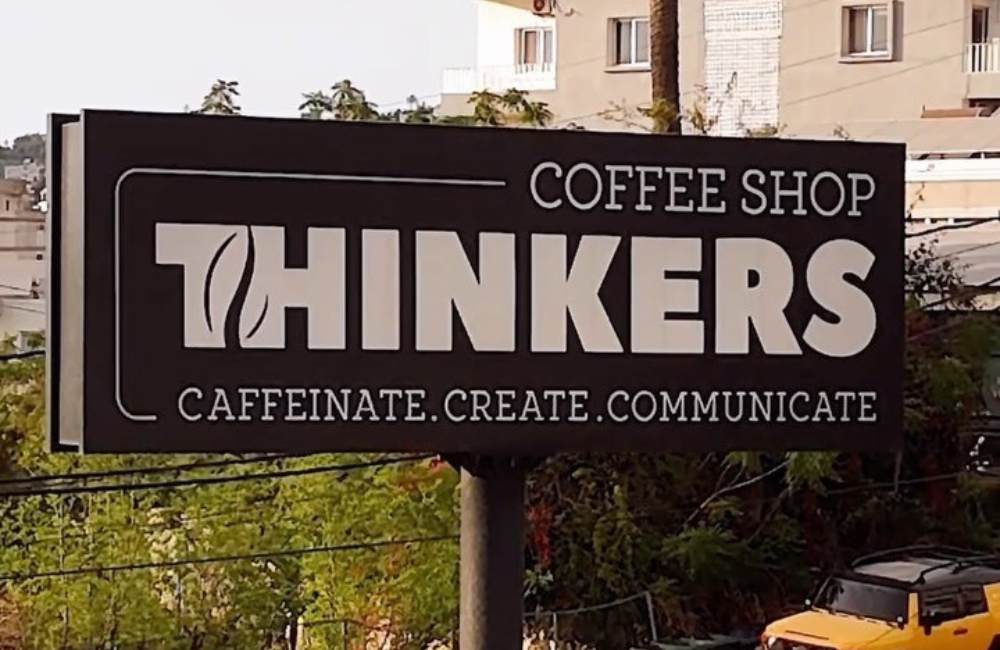 Outdoor Thinkers Coffee Shop sign mounted on a pole, reading "Coffee Shop Thinkers, Caffeinate. Create. Communicate." in white on black, with residential buildings and trees behind it.
