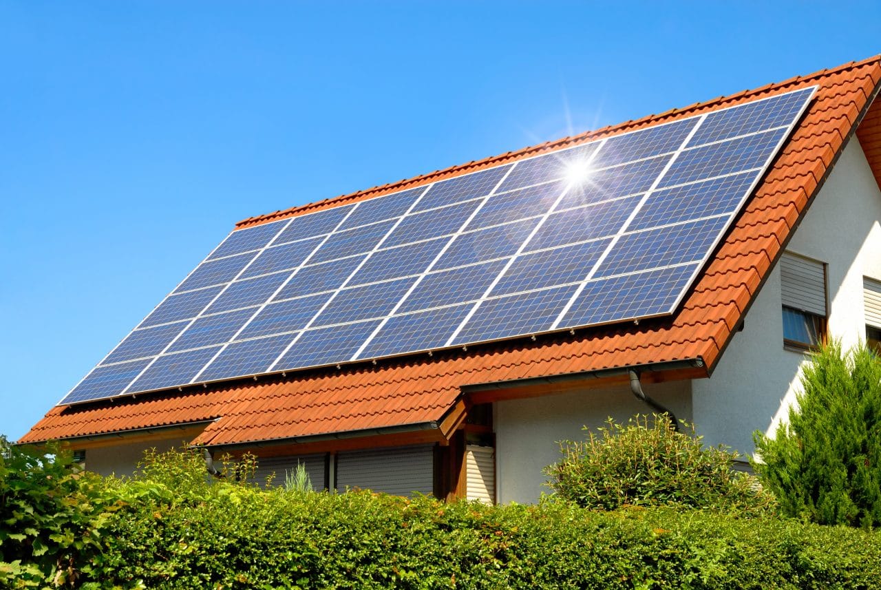Solar photovoltaic panels mounted on the red tiled roof of a modern house, with the sun flaring off the panels against a clear blue sky and green hedges in the foreground.