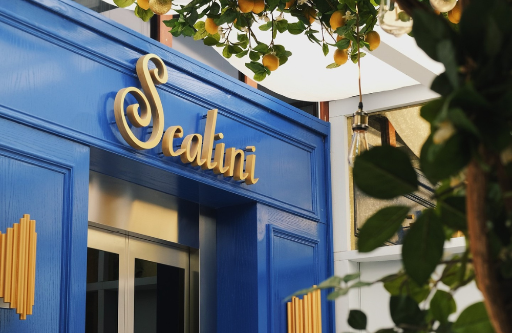 Gold cursive "Scalini" signage mounted on the restaurant's deep-blue facade, framed by an orange tree with ripe fruit and a hanging Edison-bulb light.