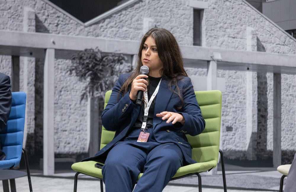 Sandra Helou sits in a green chair wearing a navy blue suit, holding a microphone as she speaks on a panel, in front of a large black-and-white photo of a stone building.