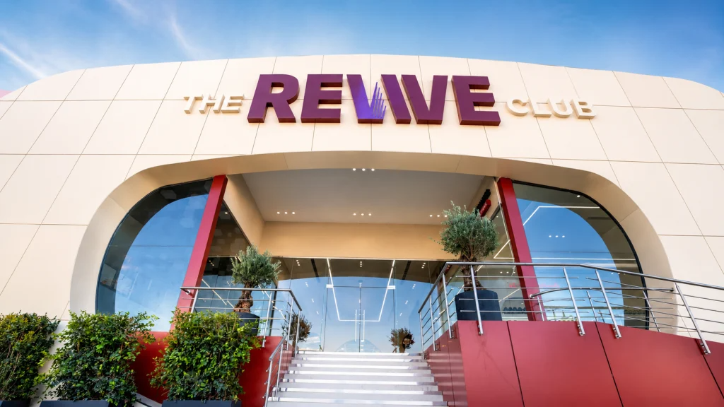 Exterior entrance of The Revive Club in Beirut, a curved cream-colored building with a purple and gold logo above a large glass archway, red accent walls, and a wide staircase flanked by olive trees under a blue sky.