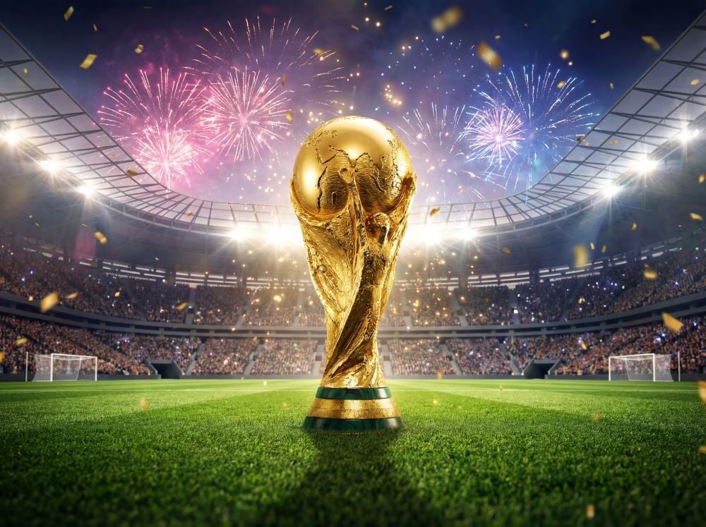 The golden FIFA World Cup trophy standing on a green stadium pitch, with fireworks and gold confetti lighting up a packed stadium at night