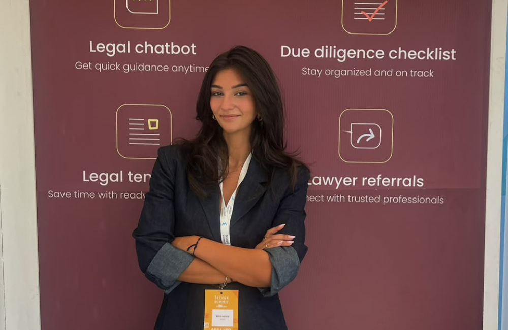 askLex founder May El Hachem stands with her arms crossed in front of a maroon askLex banner that lists the app's four features: legal chatbot, due diligence checklist, legal templates, and lawyer referrals. She wears a navy blazer and a summit speaker badge on a lanyard.