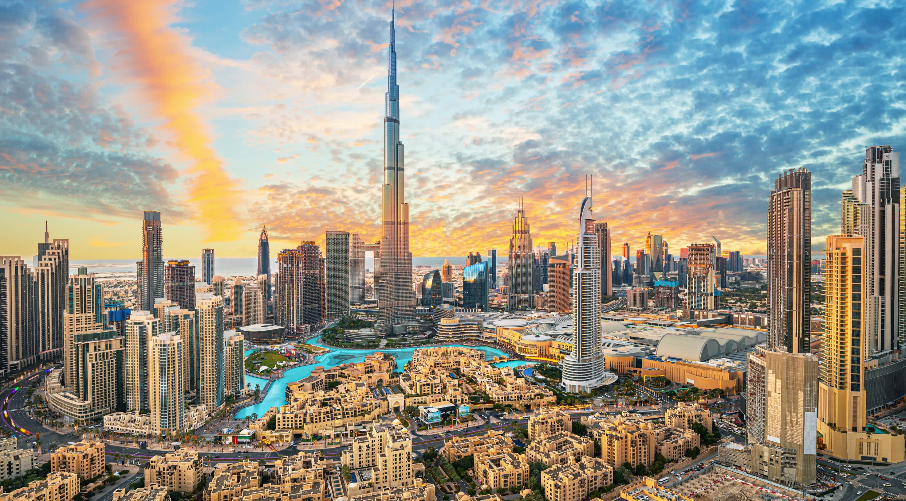 Aerial view of Downtown Dubai at sunset, with the Burj Khalifa rising above office towers, the Dubai Fountain lake and Dubai Mall