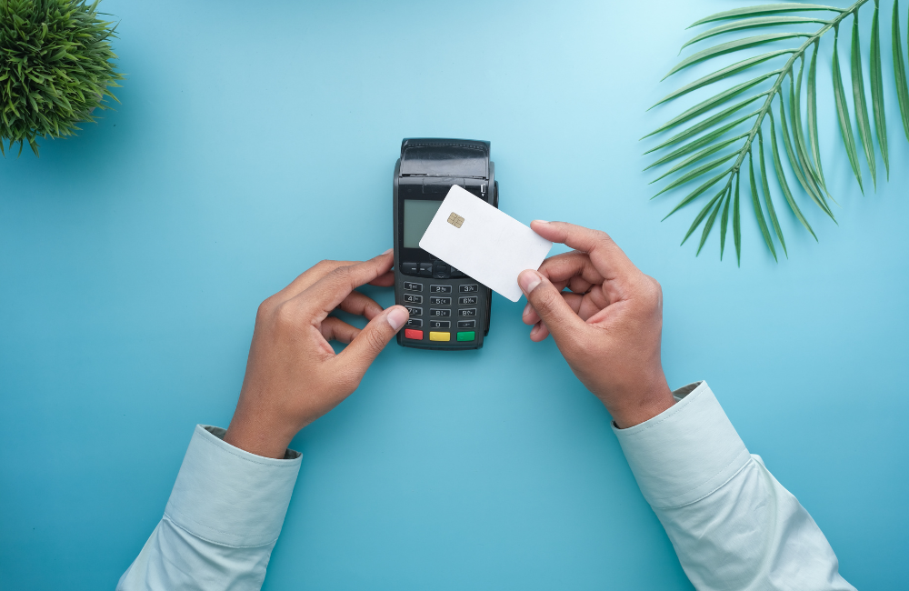 Overhead view of hands holding a blank white chip card against a black point of sale terminal on a light blue background, framed by a small plant and a palm leaf.