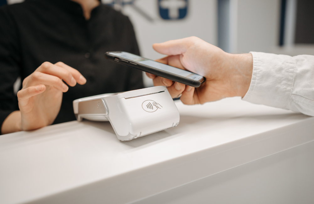 A customer holds a smartphone over a white contactless card reader on a shop counter while the cashier waits to complete the payment.