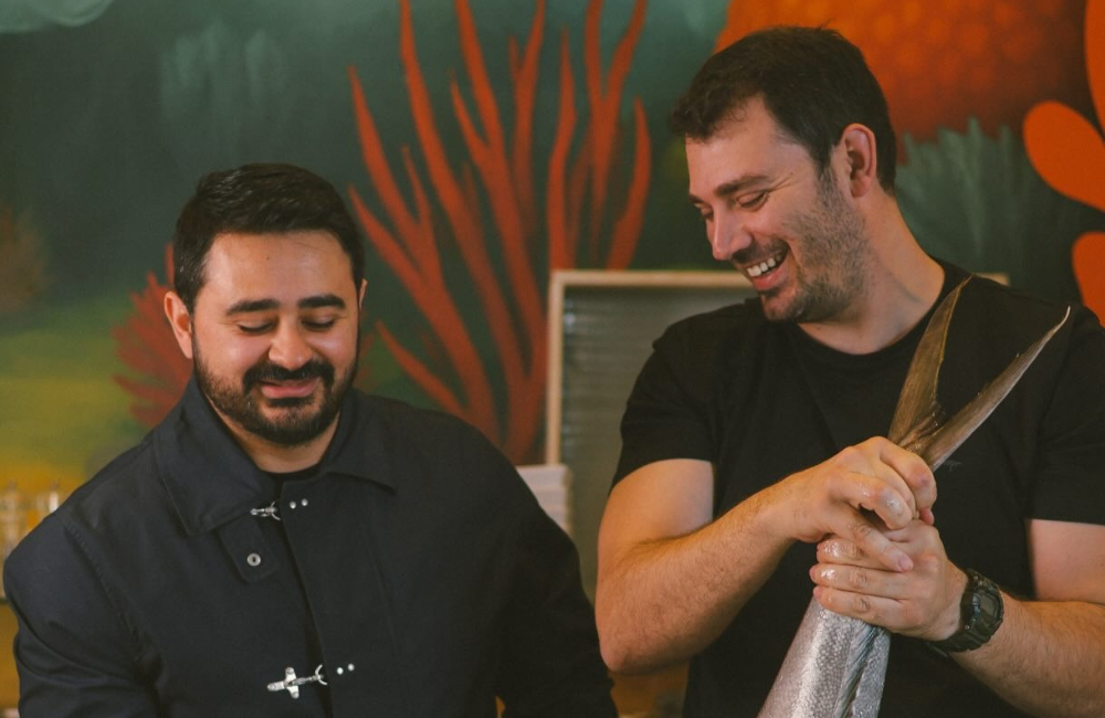 The two brothers behind Kyma laughing as one holds a fresh fish by the tail, in front of a coral mural.