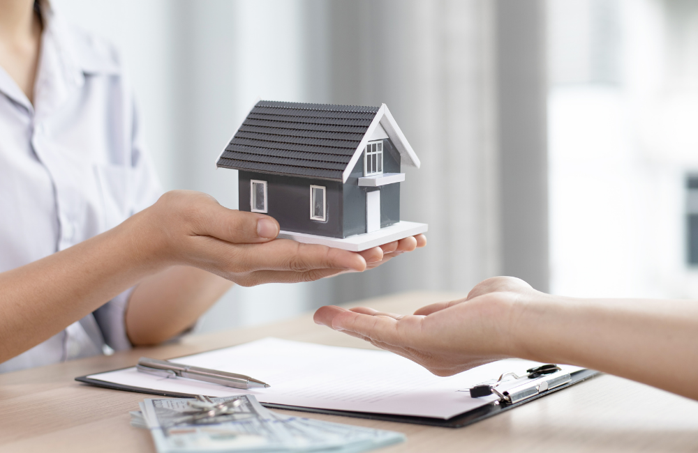 A person hands a small grey model house to another person's open palm across a desk with a clipboard, a pen, house keys and US dollar bills.