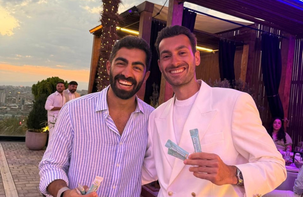 Superpowders co-founders Youssef Taher and Andy Kazanjian smile and hold packs of Superpowders electrolyte sachets at a rooftop event at sunset.