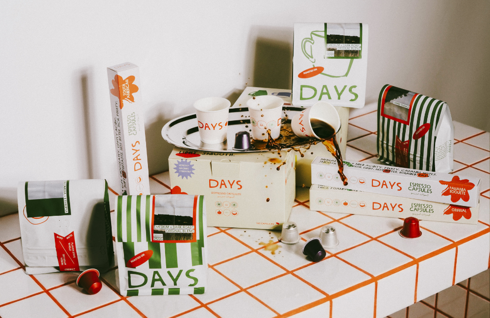 Days coffee products on a white tiled counter with orange grout: bags of coffee beans, boxes of espresso capsules and branded paper cups spilling coffee onto a silver tray.