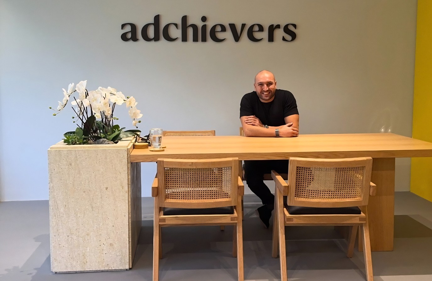 Elias Saliba, founder of Adchievers, sitting at a wooden meeting table under the Adchievers logo in the agency's office