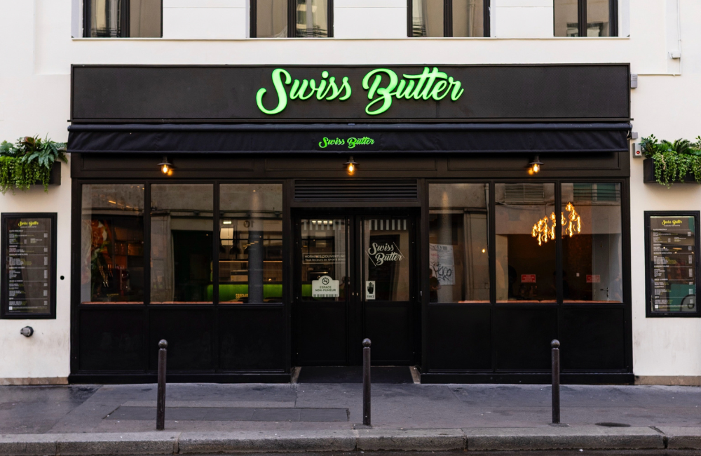 Black storefront of Swiss Butter in Paris with a green neon logo, large glass windows, and menu boards on each side of the entrance