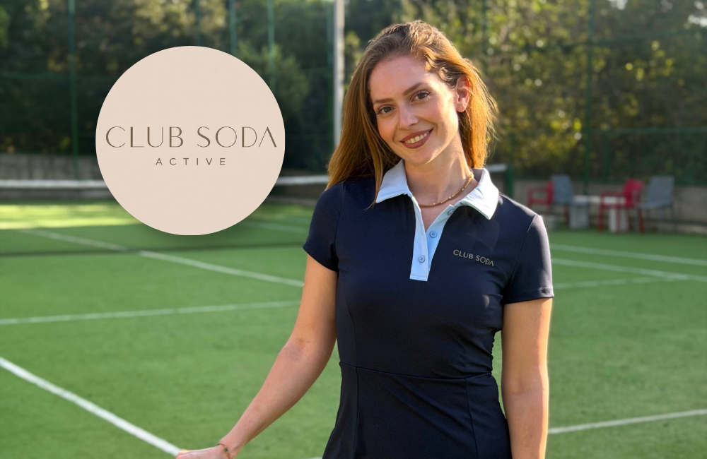 A woman smiles on a green tennis court, wearing a navy CLUB SODΛ ACTIVE tennis dress with a light blue collar. Beside her is a circular beige CLUB SODΛ ACTIVE logo.