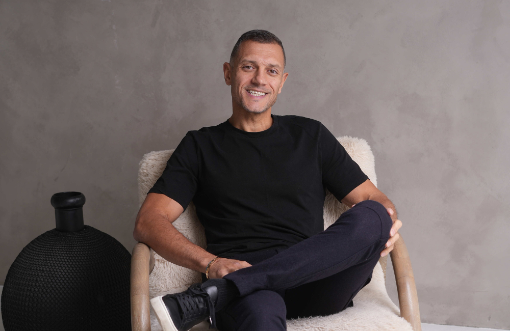 Sam Kamra, founder of Listing.ca, smiling while seated in a white sheepskin armchair, wearing a black T-shirt and dark trousers, against a grey textured wall