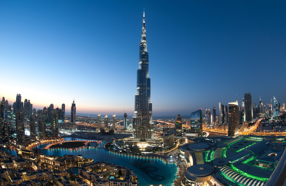 Downtown Dubai at dusk, with the illuminated Burj Khalifa rising above the Dubai Fountain lake, Dubai Mall and surrounding skyscrapers under a deep blue sky.