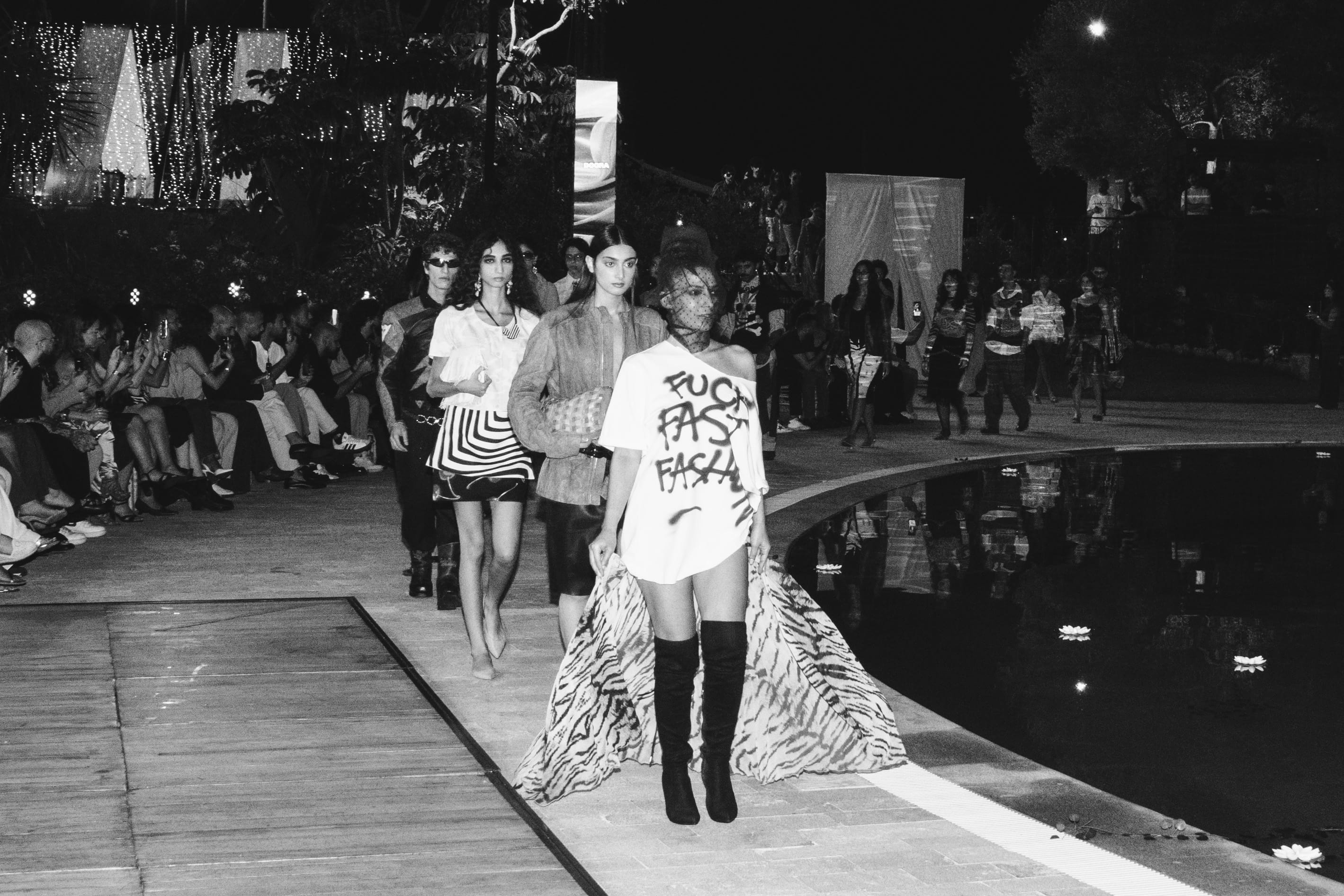 Black and white photo of models walking a poolside runway at night during DOORA’s secondhand fashion show, the lead model in a spray painted anti fast fashion T shirt, tiger print skirt and thigh high boots, with guests filming from the front row.