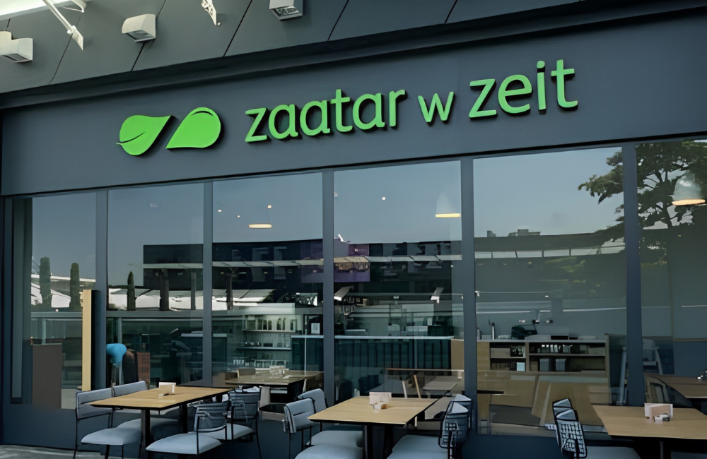 Zaatar w Zeit restaurant storefront with the green leaf logo and brand name on a dark grey facade, floor to ceiling windows, and wooden tables with grey chairs on the outdoor terrace