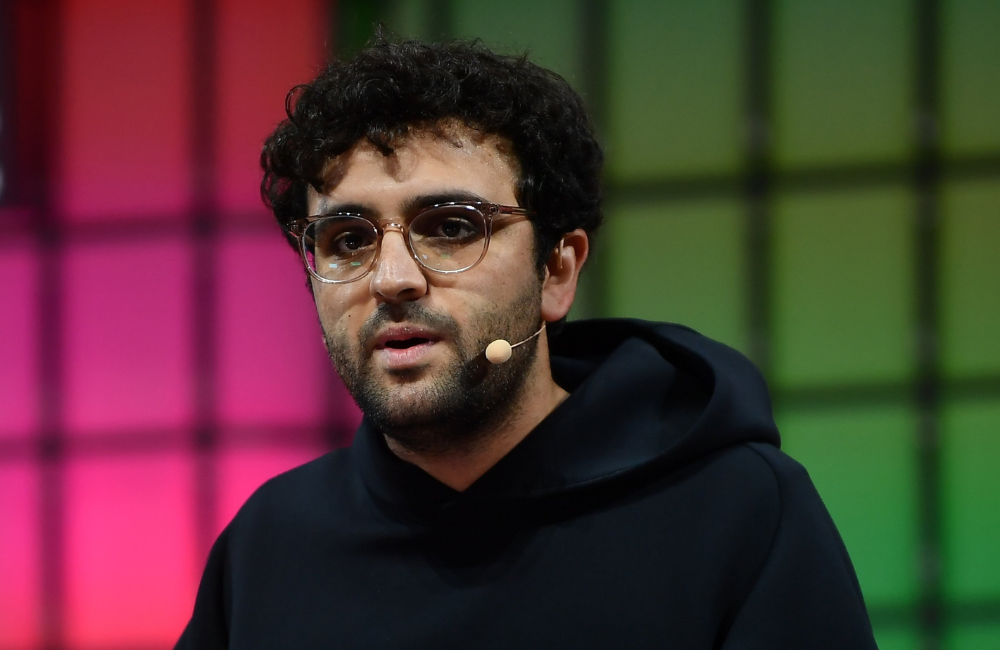 A man with curly dark hair, a beard and clear framed glasses speaks on stage wearing a black hoodie and a headset microphone, in front of a blurred pink and green backdrop.